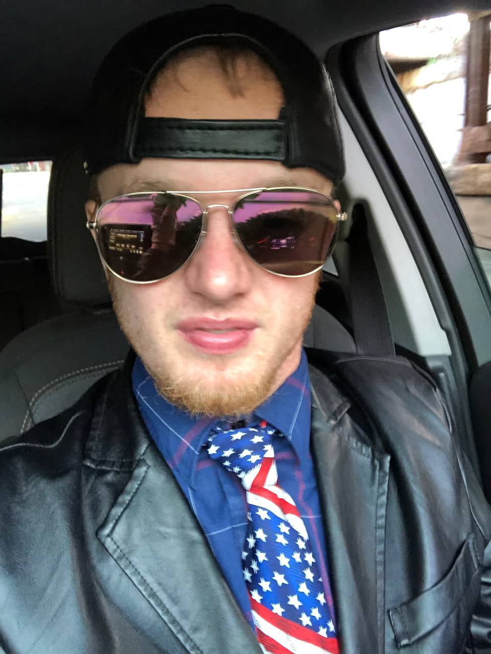 luke wenke of olean ny sitting in a vehicle in a black leather jacket and patriotic tie with a backwards baseball cap and sunglasses